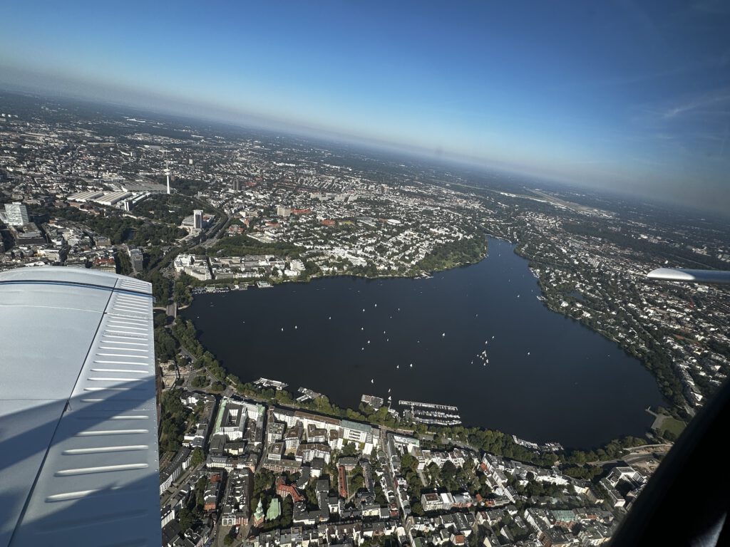 Rundflug über Hamburg mit Außenalster und Hamburger Fernsehturm. Der Himmel ist Blau und im Hintergrund ist der Hamburg Airport zu sehen.