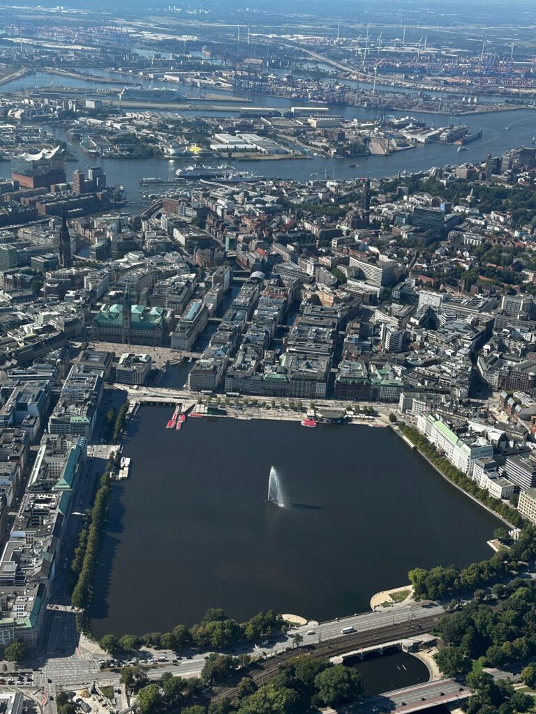 Die Hamburger Innenalster mit Springbrunnen an einem Sommertag mit blauem Himmel. Im Hintergrund ist die Elbe mit AIDA Kreuzfahrtschiff zu sehen. An der Alster sieht man während des Rundfluges den Jungfernstieg.