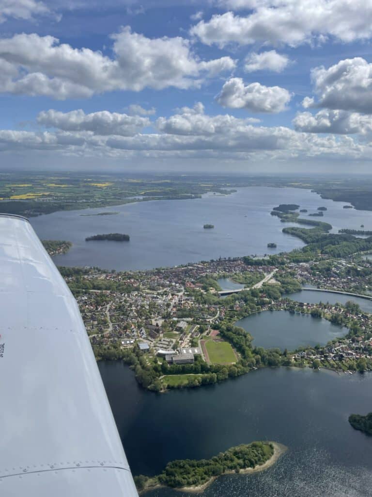 Der Große Plöner See auf einem Rundflug. Malente und Plön mit Schloss sind zu sehen. Der Himmel ist blau und zum Teil mit weißen Wolken bedeckt.